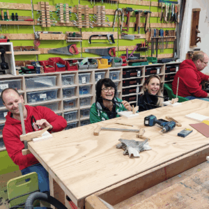 REWE-Führungskräfte sägen lachend Holzbuchstaben-Aufsteller mit der Laubsäge im Schreinerei-Teambuilding.