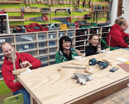 Rewe Teambuilding Führungskräfteworkshop Spaß und Kennenlernen REWE-Führungskräfte sägen lachend Holzbuchstaben-Aufsteller mit der Laubsäge im Schreinerei-Teambuilding.